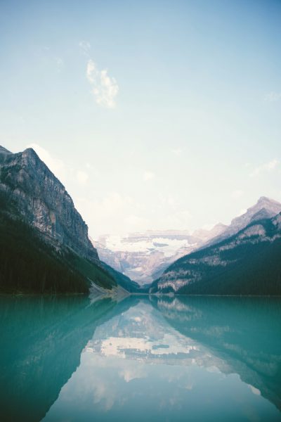 Lake Louise, Canada, landscape photography, soft tones, clear sky, reflection of the mountains in the water, shot on a film camera, wide angle shot, natural light, tranquil mood, blue and green color palette, high resolution, highly detailed. --ar 85:128