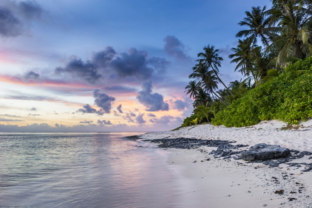 A serene beach in the Maldives at sunset, with palm trees and white sand, overlooking an island. The sky is painted with hues of pink, orange, blue, and purple as clouds gently float. In front stands a small rocky outcrop on which lush green vegetation grows. A gentle breeze rustles through the leaves creating calm waves that dance along the shoreline. This scene captures the tranquility and beauty of nature’s embrace. –ar 128:85