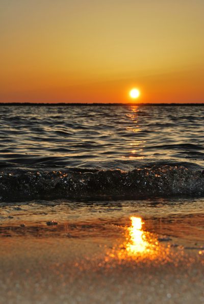 The setting sun shines on the sea, reflecting golden light in the water and shimmering waves rolling along with it. The beach is covered by fine sand that reflects the sunset's warm colors. In front of you stands an empty scene, surrounded by gentle ripples under the orange sky., Real photography, high definition details --ar 85:128