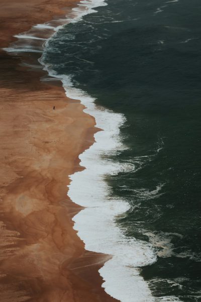 A photo of a beach from above, with waves breaking on the shore, brown sand, minimalism, in the style of dark green and amber tones, soft light, moody. --ar 85:128