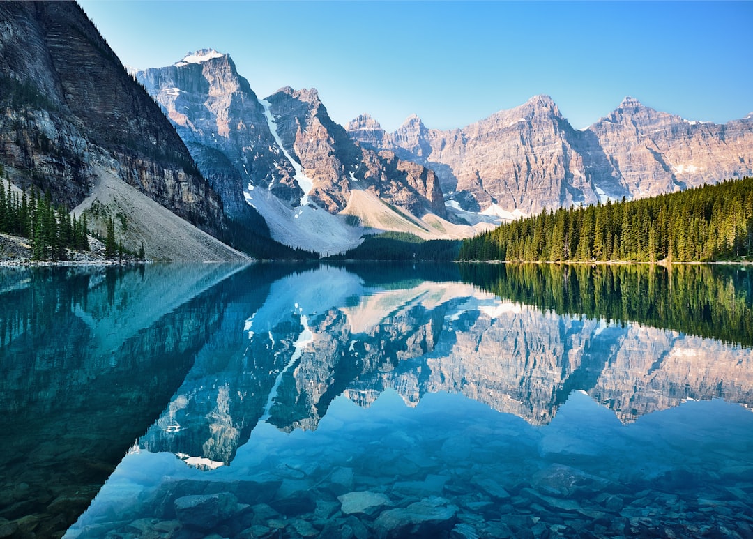 A stunning photograph of the picturesque Moraine Lake in Canada, reflecting crystal clear waters and majestic mountains. The scene captures the serene beauty of nature with towering peaks standing tall against a blue sky backdrop. The photograph was taken in the style of [Ansel Adams](https://goo.gl/search?artist%20Ansel%20Adams), known for his black-and-white landscape photographs of American national parks and wilderness areas. –ar 128:91