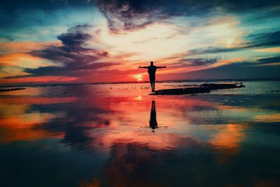 A person standing on the beach with outstretched arms, watching the sunset, with a reflection in the water, the silhouette of the figure against a colorful sky, creating a serene and peaceful atmosphere, vibrant colors, in a wideangle shot, taken with a Nikon D850 using natural lighting. --ar 128:85