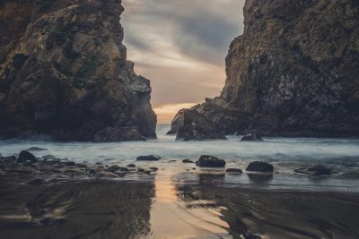 A photo of the rocky beach at Big Sur, California with an opening between two rocks that creates a small archway leading to water and sand on either side of it, cloudy sky, evening light, cinematic, shot in the style of an ARRIFLEX35 BL camera, photographic, realistic, depth of field. --ar 128:85