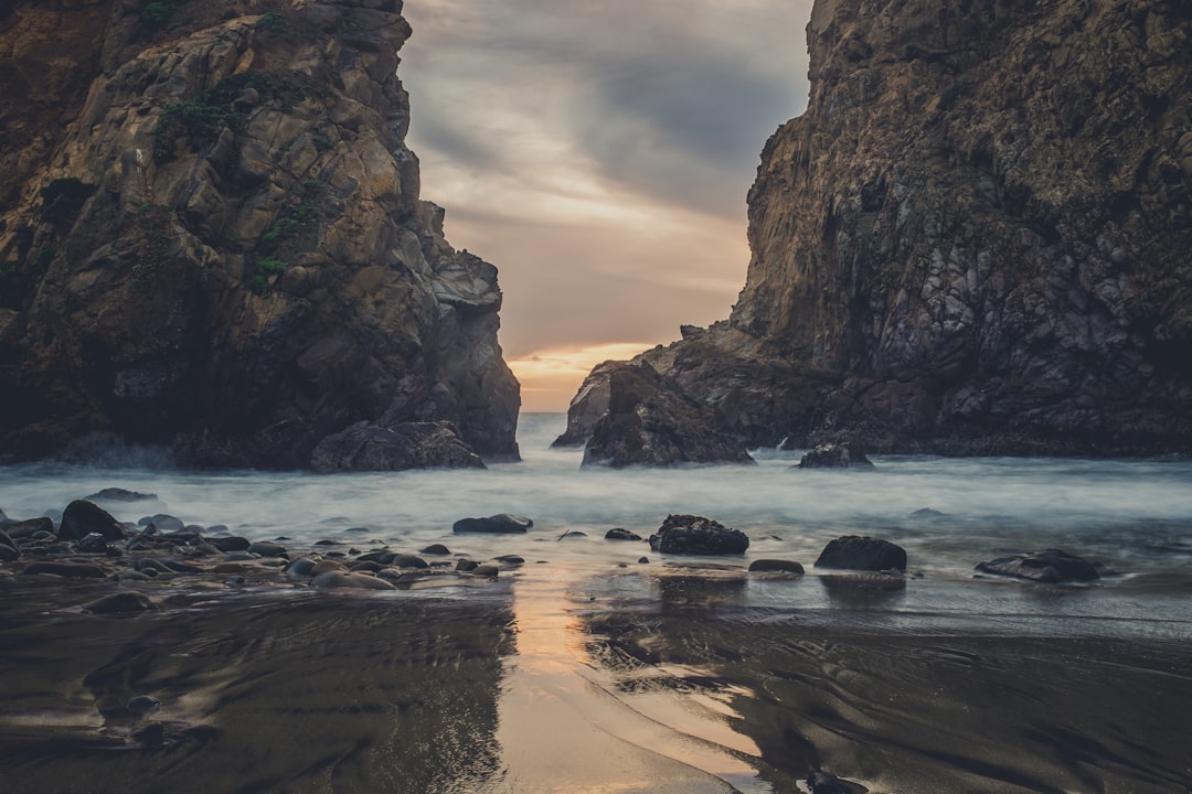 A photo of the rocky beach at Big Sur, California with an opening between two rocks that creates a small archway leading to water and sand on either side of it, cloudy sky, evening light, cinematic, shot in the style of an ARRIFLEX35 BL camera, photographic, realistic, depth of field. –ar 128:85