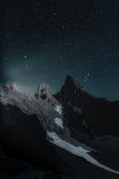 Realistic photography of mountain peaks at night, stars in the sky, dark and mysterious atmosphere, high contrast between light from behind and darkness below, sharp details on snow-covered rocks, moonlight casting long shadows over the scene, creating an ethereal feeling of wonder. --ar 85:128