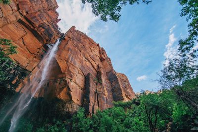 low angle photo of a waterfall flowing down the side of a red rock cliff in zion national park, with lush green trees and a blue sky, in the style of unsplash photography --ar 128:85