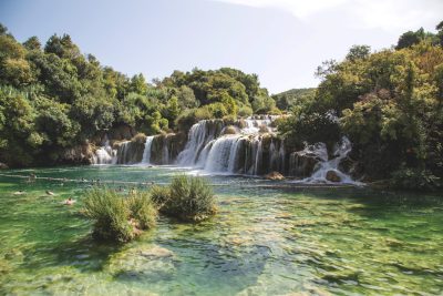 A wide shot of the cascading waterfalls at Krka National Park in Croatia, surrounded by lush greenery and crystal clear waters with people swimming or sitting on stones. The sky is a clear blue, creating a picturesque scene. Shot during midday to capture natural light. A Canon EOS camera with an aperture set at f/5.6 for sharpness and bokeh effect. Softbox lighting was used to highlight details and create depth in the frame. In the style of hyperrealistic photography. --ar 128:85