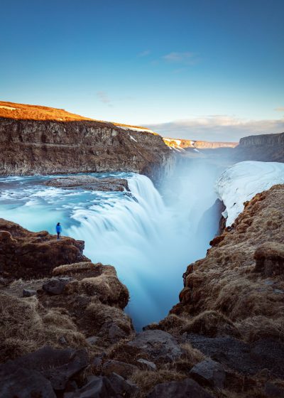 Photo of Gullfoss waterfall in Iceland, with blue sky and man standing on the edge looking at it, high resolution photography, dslr camera, travel magazine photo, awardwinning photograph, wide angle lens, long exposure time, golden hour lighting, --ar 91:128