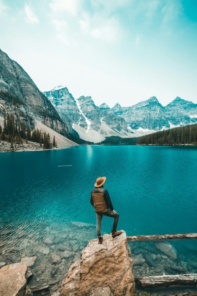 A man standing on the edge of Lake Moraine in Alberta, Canada with beautiful blue water and mountains behind him. He is wearing outdoor  and a hat. The sky above his head has light white clouds and there is some forest around it. --ar 85:128