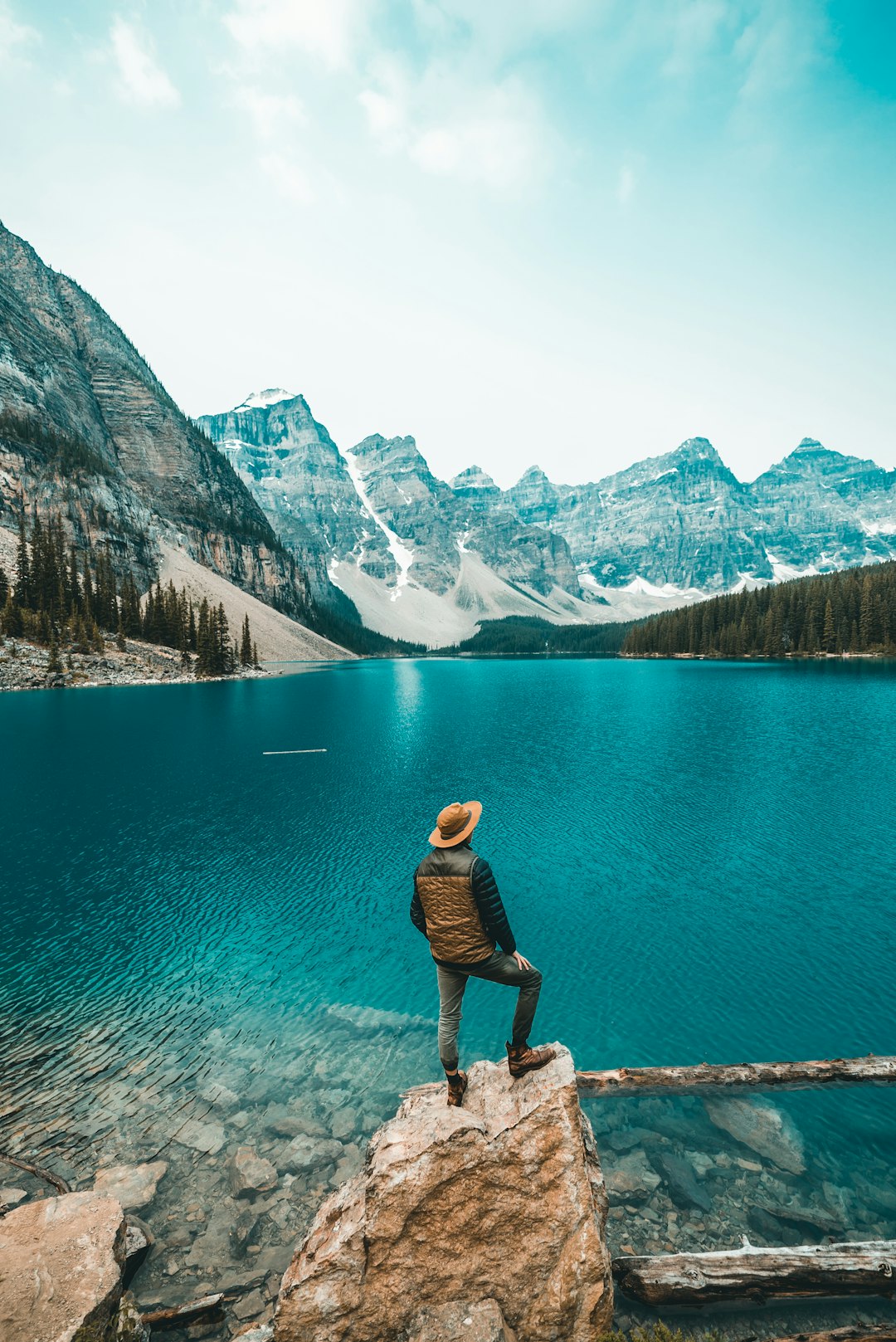 A man standing on the edge of Lake Moraine in Alberta, Canada with beautiful blue water and mountains behind him. He is wearing outdoor  and a hat. The sky above his head has light white clouds and there is some forest around it. –ar 85:128