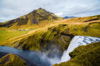 Beautiful view of Skogafoss waterfall in Iceland, landscape photography, wide angle lens, green grassy hills, water flowing down the mountain and into river below, sky with white clouds, small village on distant hillside, sunlight shining through the mist from behind mountains, cinematic photography style, photo taken in the style of Sony Alpha A7 III camera with a Sony FE2485mm f/3.64 O.I.S., sharp focus, high resolution --ar 128:85