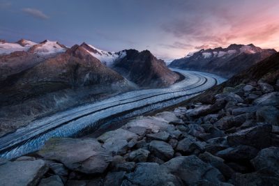 In the Swiss Alps, at sunset, the Aletchigee glacial river flows between snowcapped mountains, creating an awe-inspiring natural landscape. The image captures the vastness of nature with its wide-angle lens and a cinematic feel, highlighting the contrast in colors from blue to pink as sunlight sets on rocks and ice. The camera angle is high above ground, offering a panoramic view that highlights both distant peaks and close-up details like water splashes or rocks along the stream's path. --ar 128:85