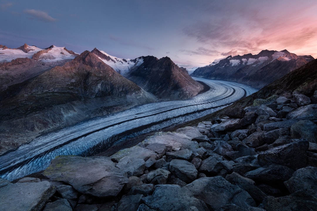 In the Swiss Alps, at sunset, the Aletchigee glacial river flows between snowcapped mountains, creating an awe-inspiring natural landscape. The image captures the vastness of nature with its wide-angle lens and a cinematic feel, highlighting the contrast in colors from blue to pink as sunlight sets on rocks and ice. The camera angle is high above ground, offering a panoramic view that highlights both distant peaks and close-up details like water splashes or rocks along the stream’s path. –ar 128:85