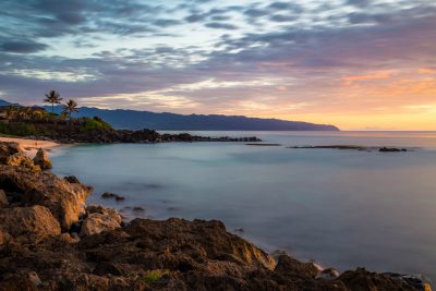 Long exposure shot of the shore at sunset in Hawaii, with rocks and mountains visible on the distant horizon. The calm sea reflects pastel colors from the sky. In front is an empty beach with small waves lapping against it. A lone palm tree stands to one side, adding contrast between land and water. There is a hint of human presence with houses seen far away along the mountainous coastline, in the style of an empty beach scene. --ar 128:85