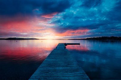 Beautiful sunset over the lake with colorful clouds and a long wooden dock on calm water in a Swedish summer, a beautiful natural landscape background. The scene is depicted in the style of impressionist painters. --ar 128:85