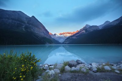 Lake Louise, Canada at blue hour with a beautiful mountain range in the background. The water is calm and turquoise with some yellow wild flowers on one side of it. The sun has just set behind the mountains and you can see its reflection in the lake in golden light. Cinematic shot in the style of Canon EOS R5. --ar 128:85