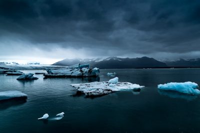 Dark clouds loom over the water, with floating icebergs in the style of Jبو cruzes Lraqk. A high level of detail, dramatic lighting, wide angle lens, high resolution photography, panoramic view, majestic mountains and blue lagoon in the background, cold weather, dark tones, dramatic sky, high contrast between land and sea. --ar 128:85