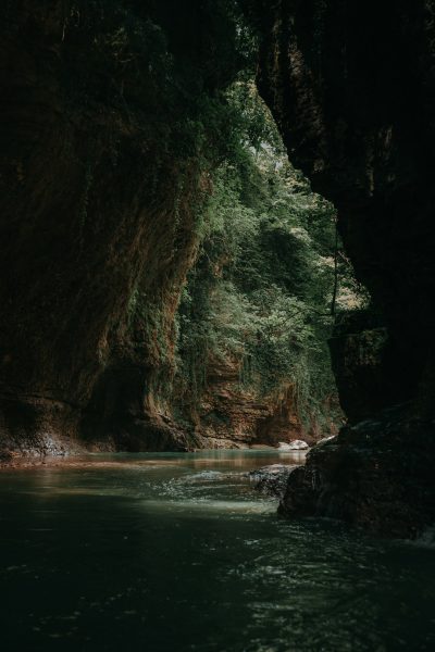 In the dark green cave, there is an underground river flowing through it. The water flows slowly and gently in front of you. On both sides of the cliff walls on either side, dense vegetation grows. In summer, it's very hot outside. Shot with Sony A7R IV camera, 2435mm f/8 lens, aperture at F6, shutter speed around 0.9 s, and ISO set to 160 for a soft light effect. This photo was taken by photographer James White.,,in --ar 85:128