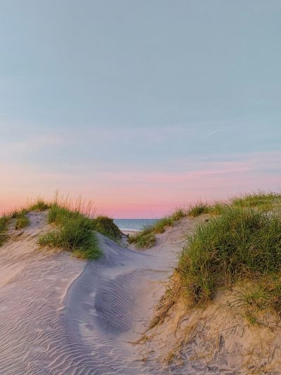 A photo of the dunes at Hamptons beach in Long Island, with a pink and blue sky, with grassy areas on both sides, sand hills, a calm sea at dusk, in the style of dummy. --ar 3:4