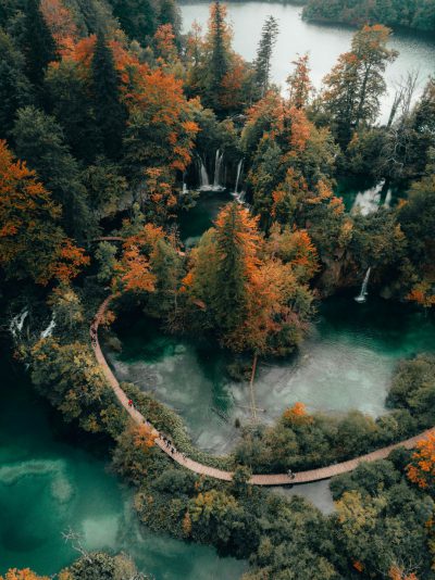 Photo of Plitvice National Park in Croatia, bird's eye view from a high angle shot taken with a drone, autumn colors of vibrant green and orange foliage with blue water, people walking along wooden pathways and a waterfall cascading into the lake below, a serene atmosphere with lush forest surrounding the park, high resolution with detailed texture in the style of drone photography. --ar 95:128