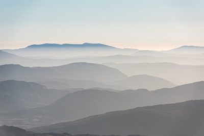 A photograph of the top view of layered mountains, with a light blue sky in the background. The foreground features a misty valley, creating an ethereal and dreamlike atmosphere. This scene captures the serene beauty of nature's landscapes. Taken by Canon EOS R5 camera with 2470mm f/8 lens at ISO 64, shutter speed 3s, soft focus on distant hills. --ar 128:85