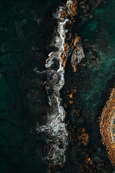 Aerial photography of the sea shows an island in front with rocks on both sides and waves crashing against them. The dark green water reflects light orange stones, creating high contrast between colors. High definition details captured with a wide-angle lens show the natural environment with flowing water and waves splashing, creating a mysterious atmosphere. Colorful dark tones add mystery to it, in the style of impressionist painters. --ar 85:128