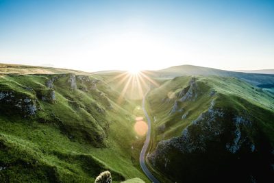 A wide-angle photo of the sun setting over rugged green hills with a winding road leading to it, shot on a Sony Alpha A7 III with an f/2 lens, capturing the dramatic interplay between sunlight rays and shadows in the English countryside in the style of light play. --ar 128:85