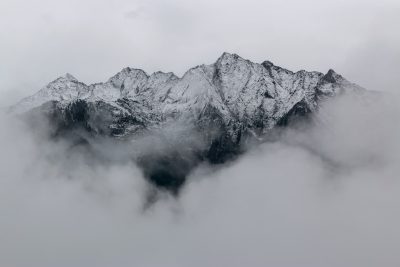 A snow-covered mountain range partially obscured by low clouds, creating an ethereal and mysterious atmosphere. The peaks stand tall against the grey sky, adding depth to the scene. Shot with a Canon EOS R5 camera using a wide-angle lens at f/8 for a dramatic effect. --ar 128:85