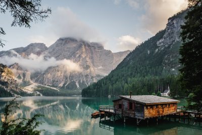 Photo of Lake Braies in the Dolomites, Italy in the style of generative AI, with a wooden house and mountain background, during summer time, depicting a beautiful natural scenery, with calm lake water, a mountain range behind, a green forest of trees, clouds at the top, capturing a natural landscape through photography, under a cloudy sky, conveying an Italy travel experience photo with an adventurous vibe, using natural light, during morning time, as a high resolution photography, --ar 128:85